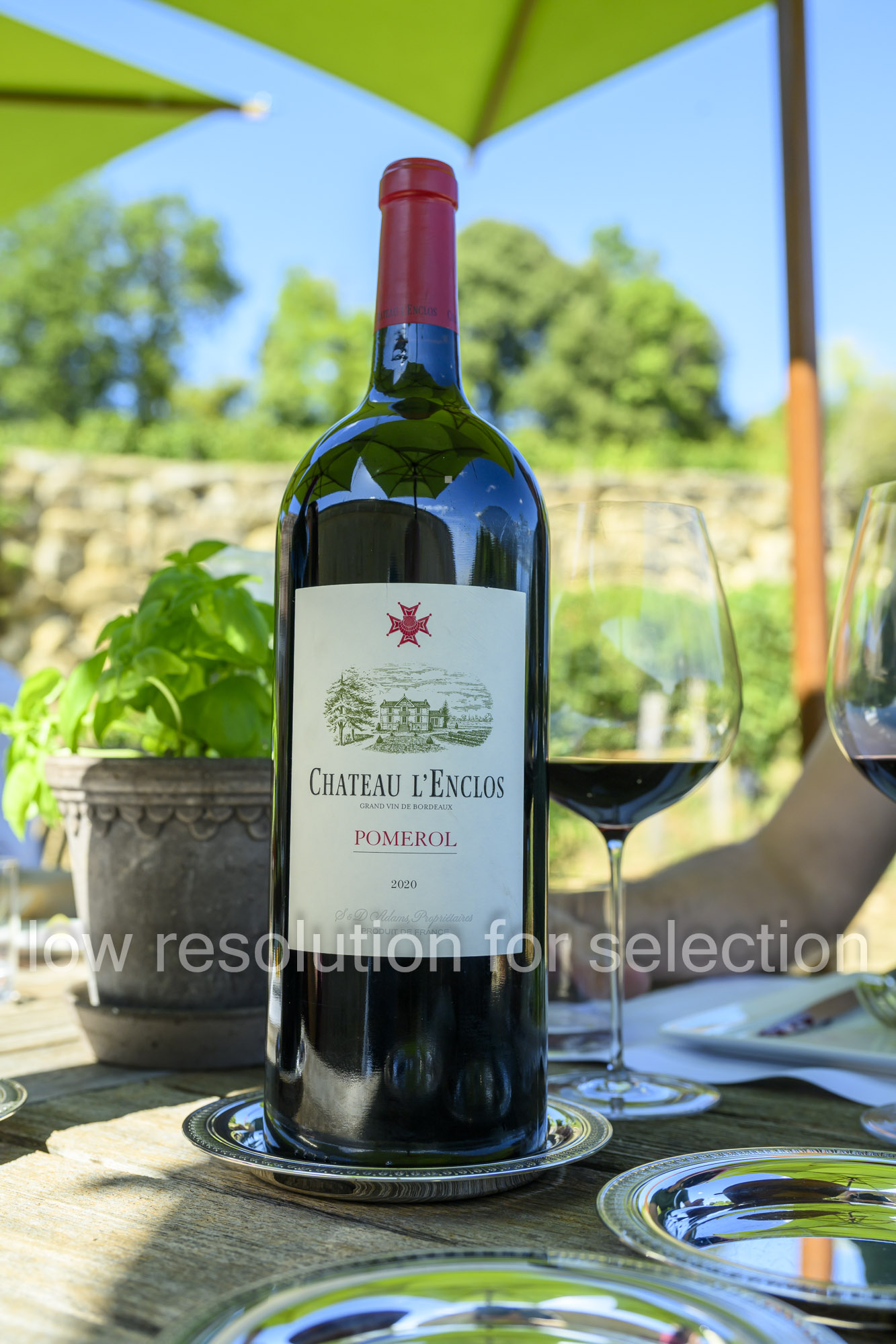 A bottle of Chateau L’Enclos Pomerol 2020 red wine standing on a wooden table next to a wine glass.