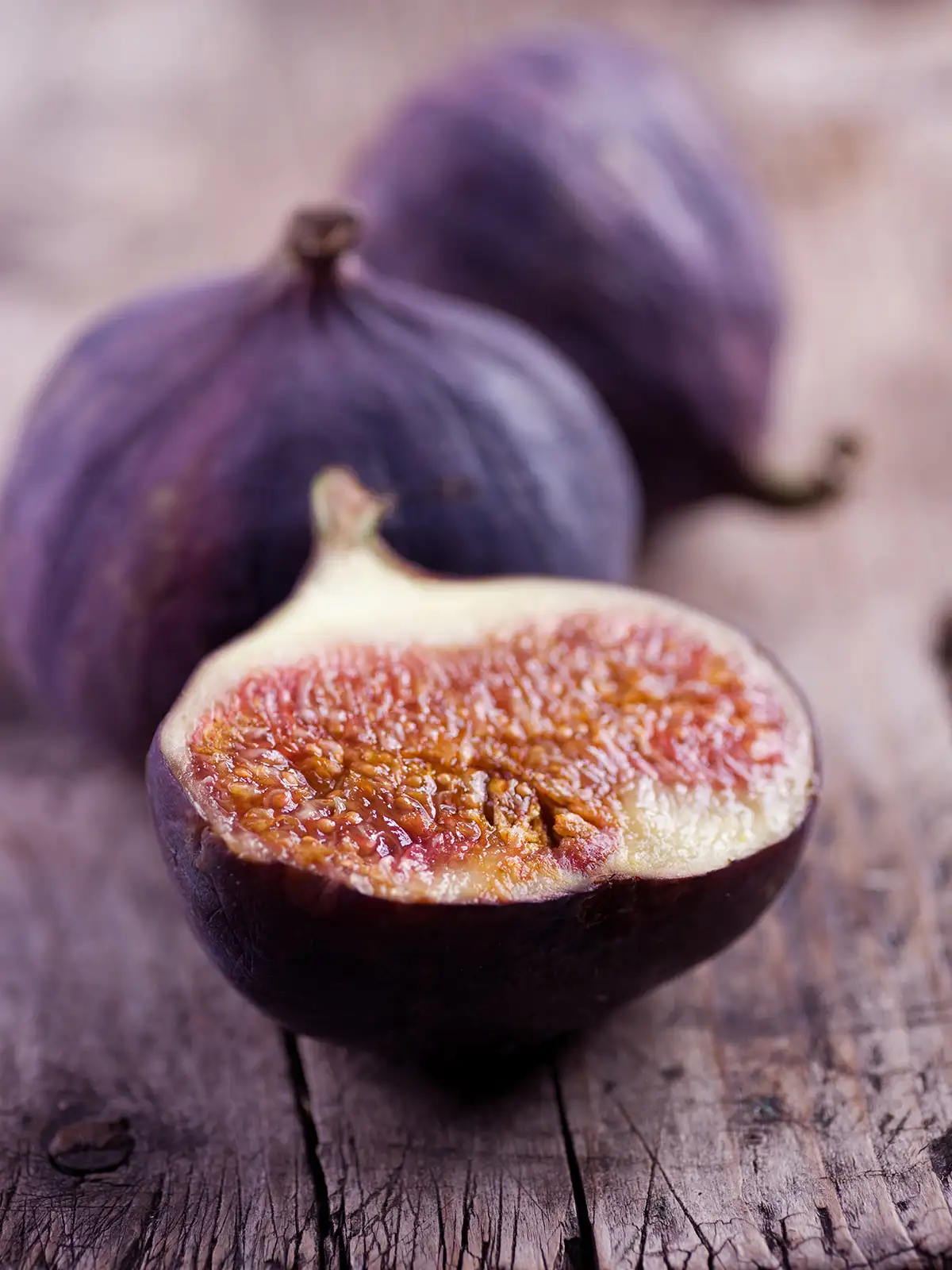 A close-up of a ripe fig cut in half, showing its pinkish interior, with two whole figs in the background on a rustic wooden surface.