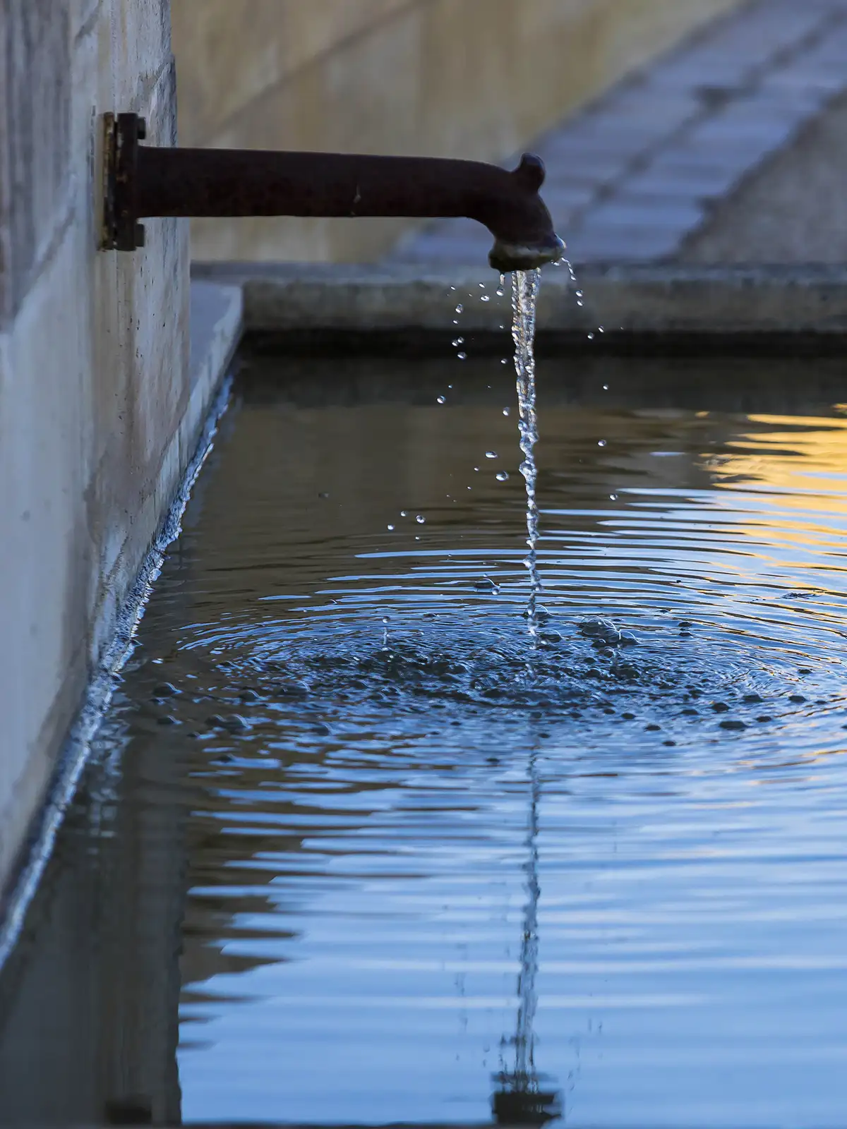 A rusted pipe extends from a wall, releasing a steady stream of water into a calm, reflective pool below.