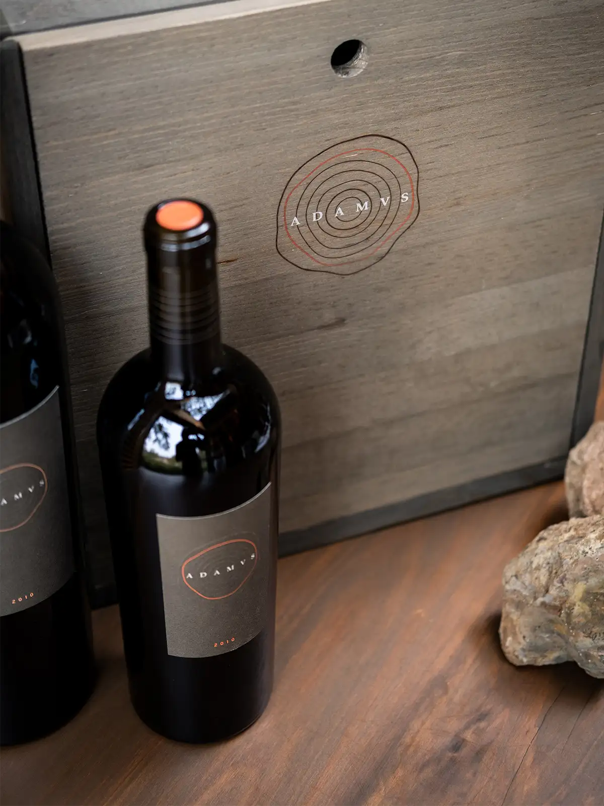 A bottle of Adamas red wine stands in front of a wooden box with the Adamas logo, next to two large rocks on a wooden surface.
