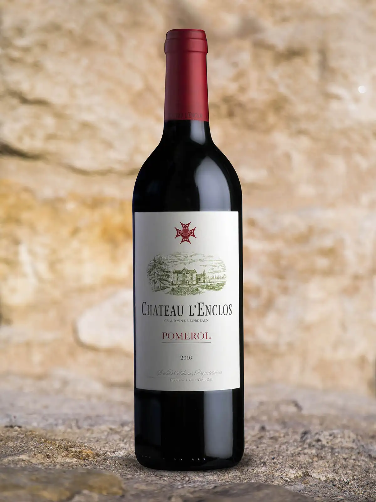 A bottle of Chateau L’Enclos Pomerol 2010 red wine stands upright on a stone surface, with a textured stone wall background.