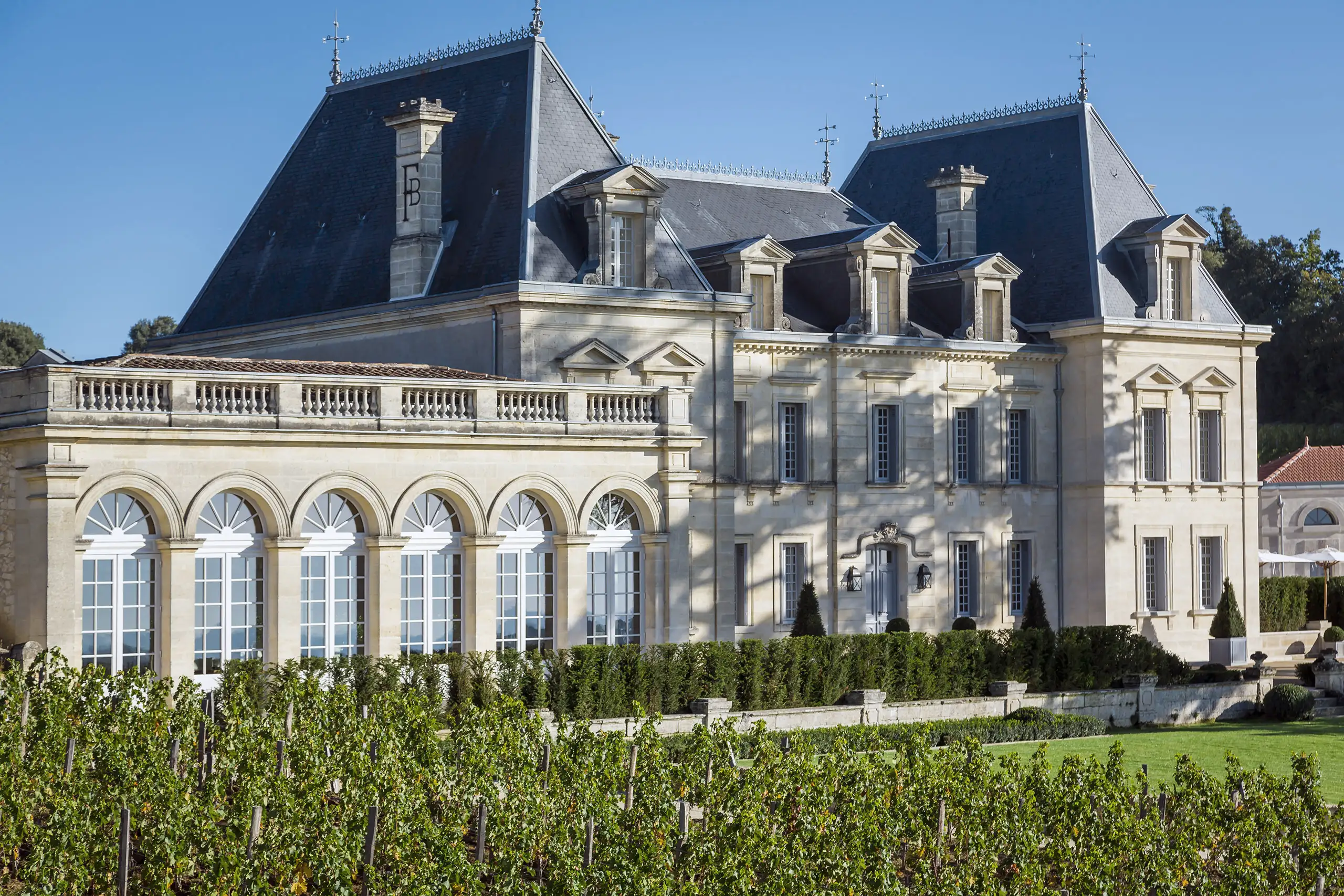 Chateau Fonplegade with mansard roofs and arched windows, surrounded by a lush green vineyard under clear skies.