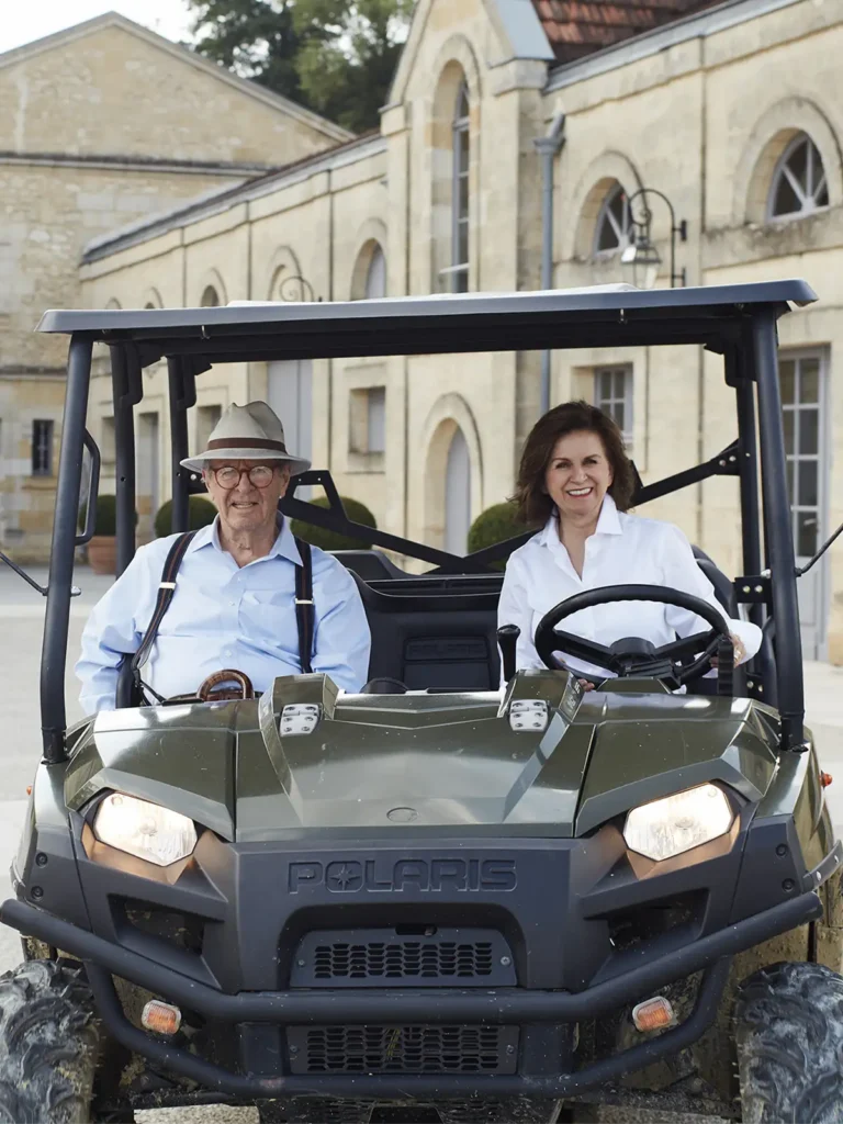 An older man and a woman sit in a Polaris utility vehicle parked in front of a stone building with arched windows.