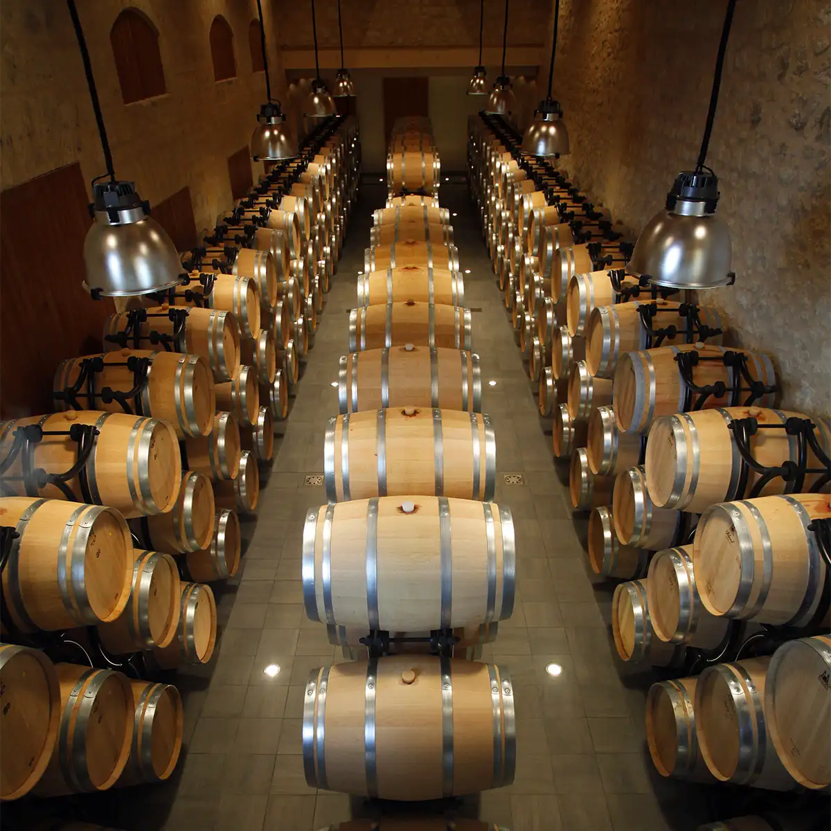 A wine cellar with rows of wooden barrels stacked on metal racks, illuminated by overhead lights, and stone walls lining the room.