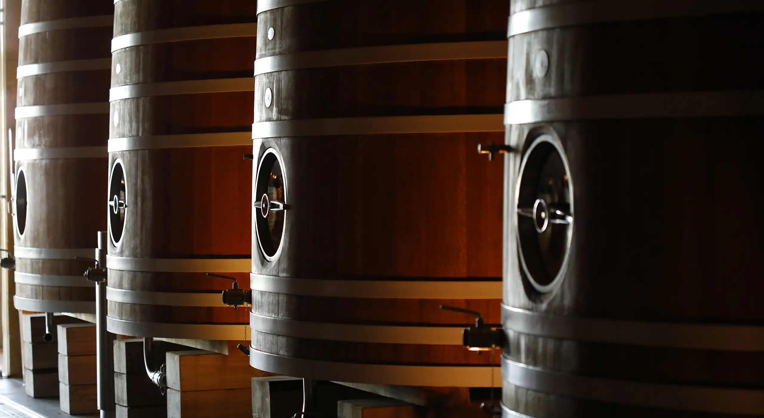 Large wooden barrels with metal bands and valves are lined up in a dimly lit area, likely for fermenting or storing beverages such as wine or beer.