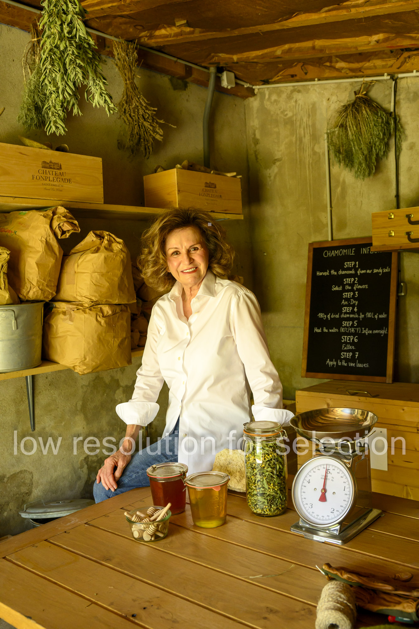 A woman in a white shirt sits at a wooden table with jars of honey, dried chamomile, and a kitchen scale, next to a chalkboard listing chamomile preparation steps.