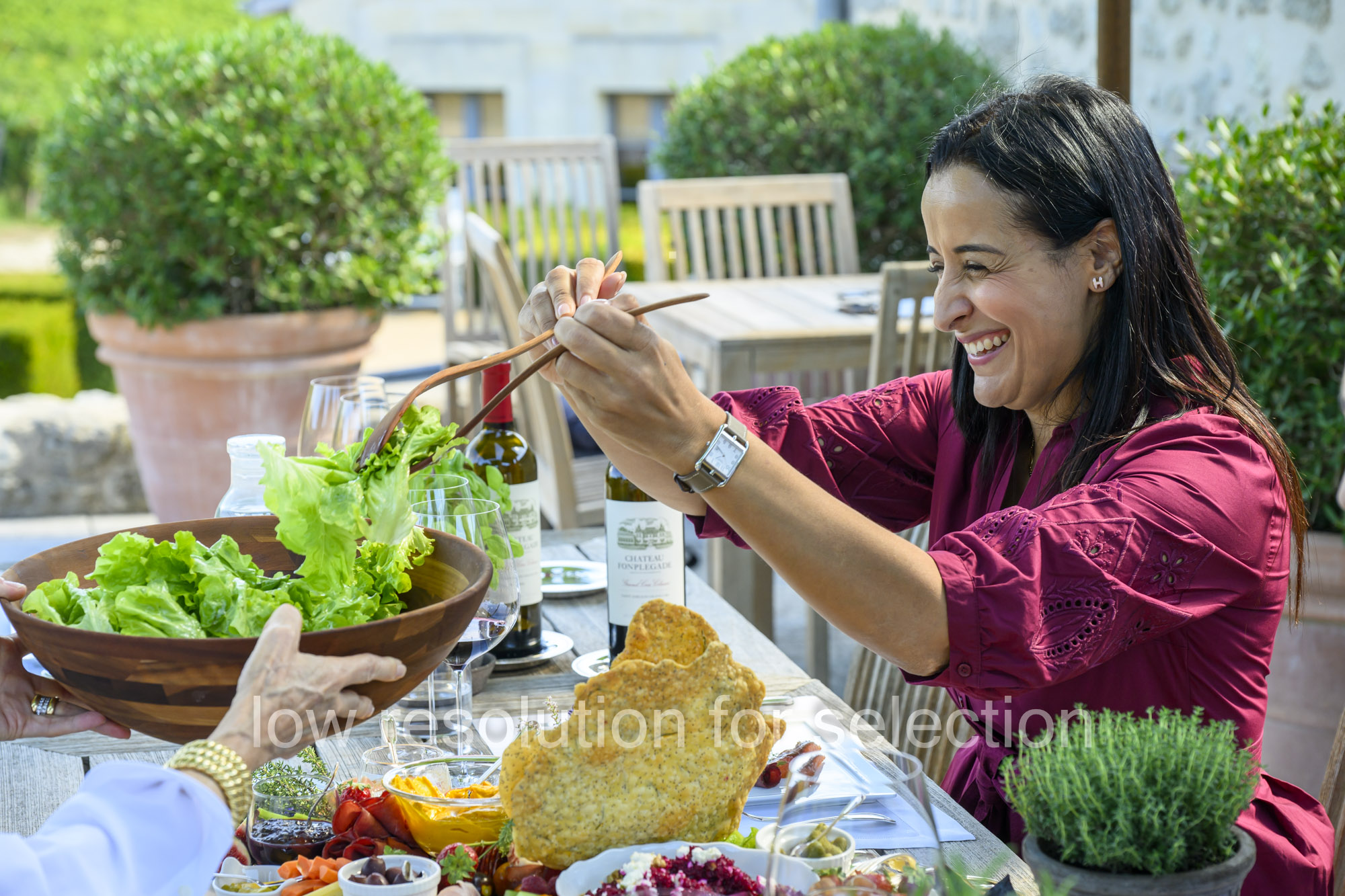 A smiling woman in a maroon dress tosses a salad with wooden serving tongs at an outdoor table.