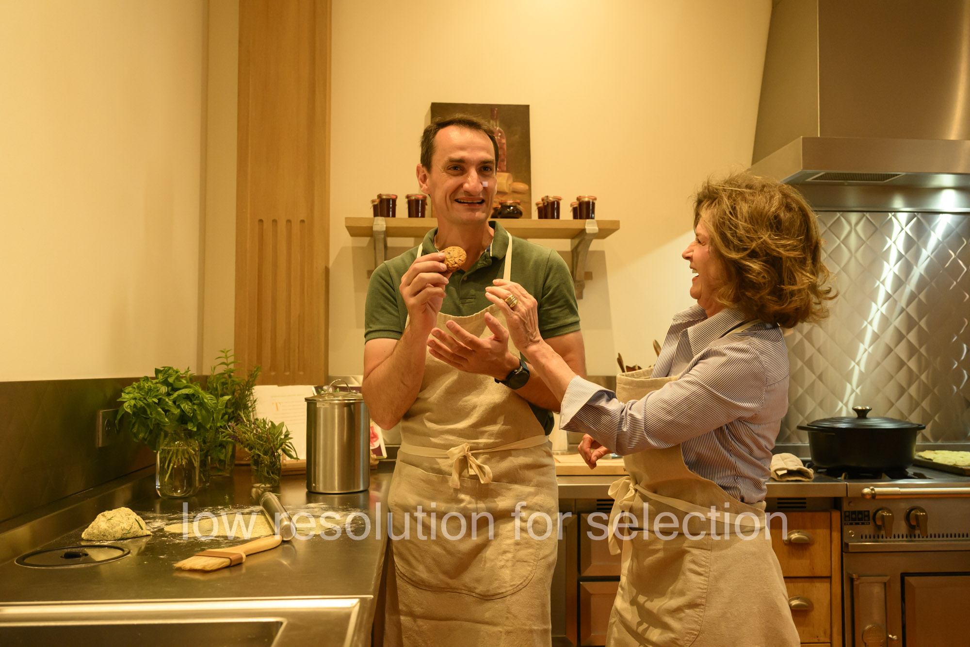A man and a woman wearing aprons in a kitchen, with the man holding up a cookie.