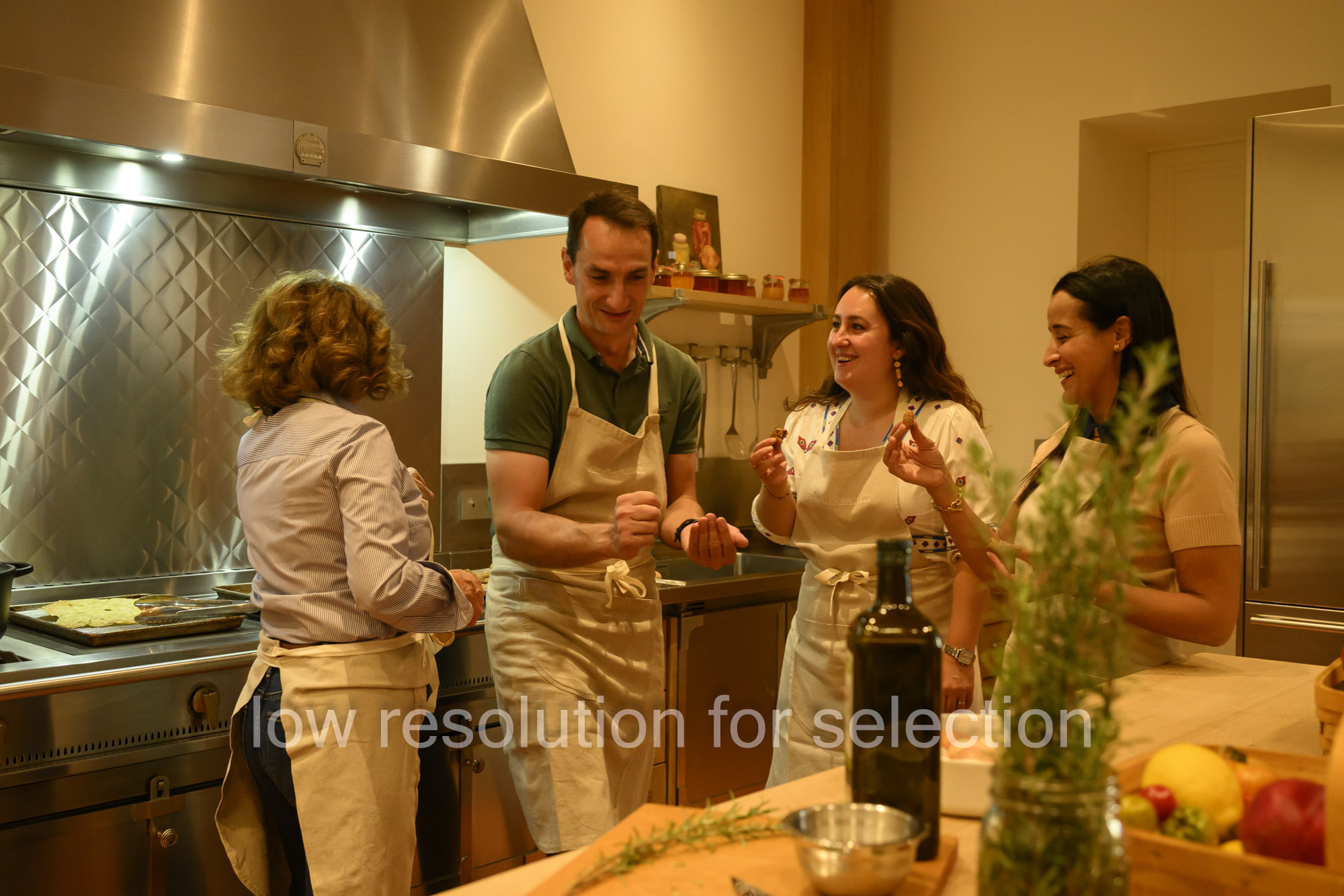 Four people in aprons stand in a kitchen, holding and tasting small pieces of food while talking.