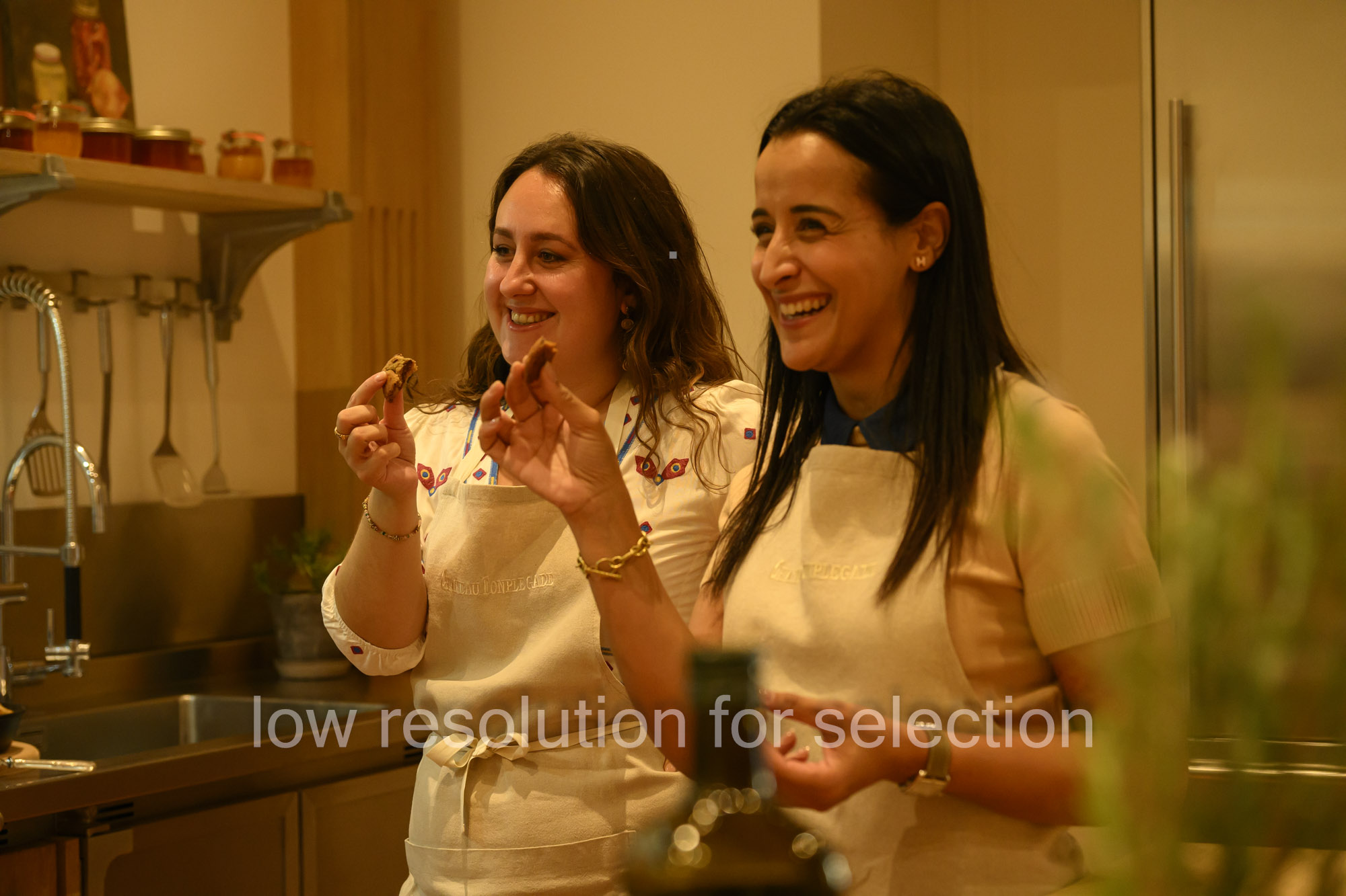 Two women wearing aprons smiling while holding and sampling food in a kitchen.