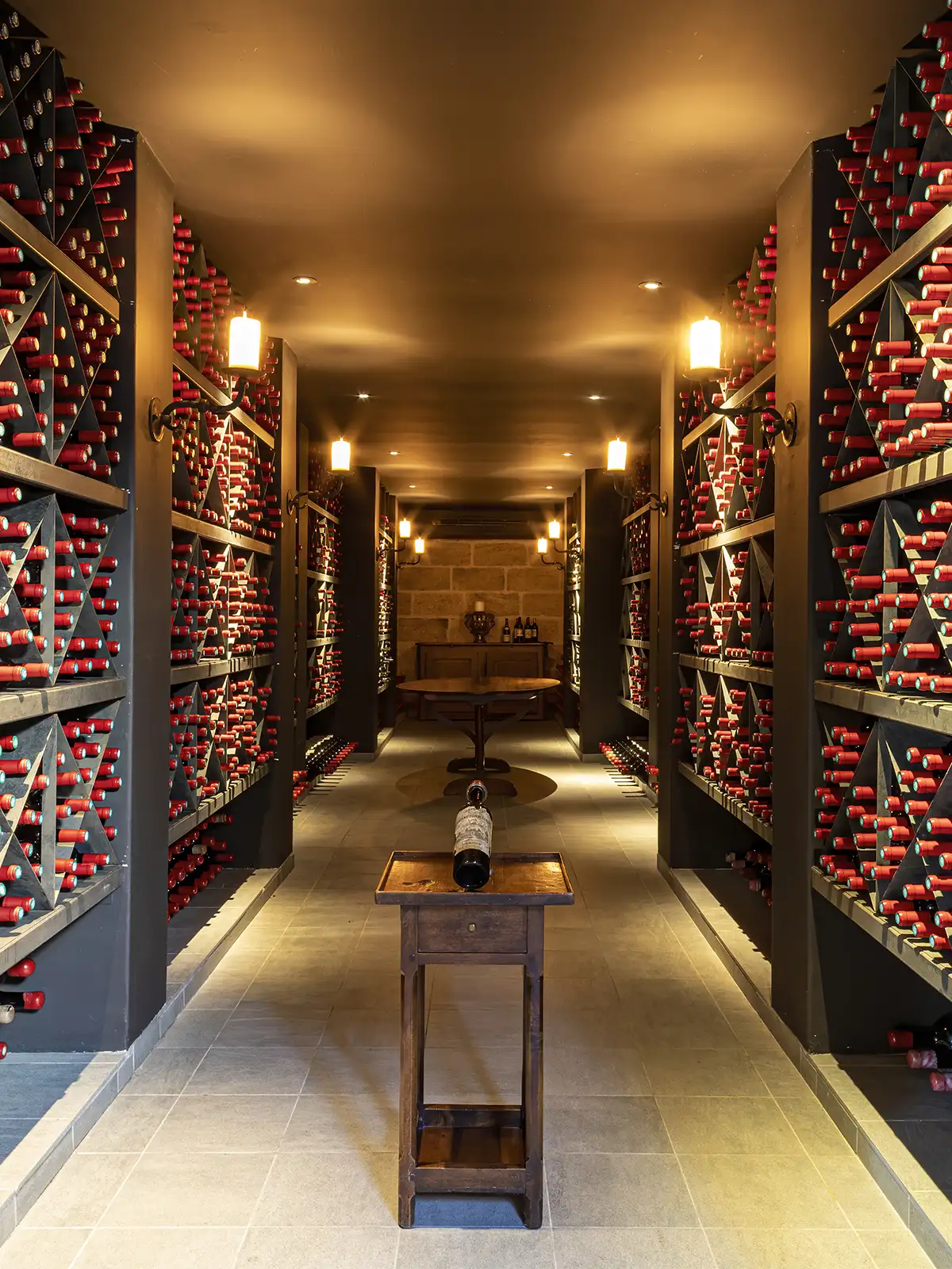 A wine cellar corridor lined with floor-to-ceiling racks of wine bottles, featuring a central pedestal table holding a single bottle.
