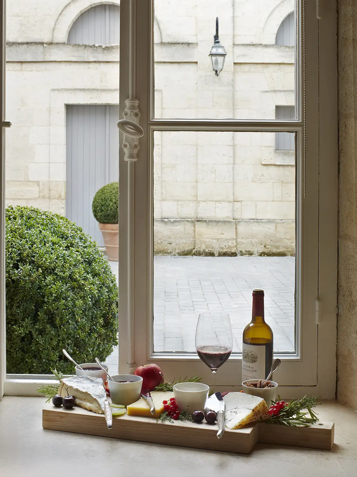 A charcuterie board with cheeses, red wine, and fruit arranged on a windowsill overlooking a stone courtyard.