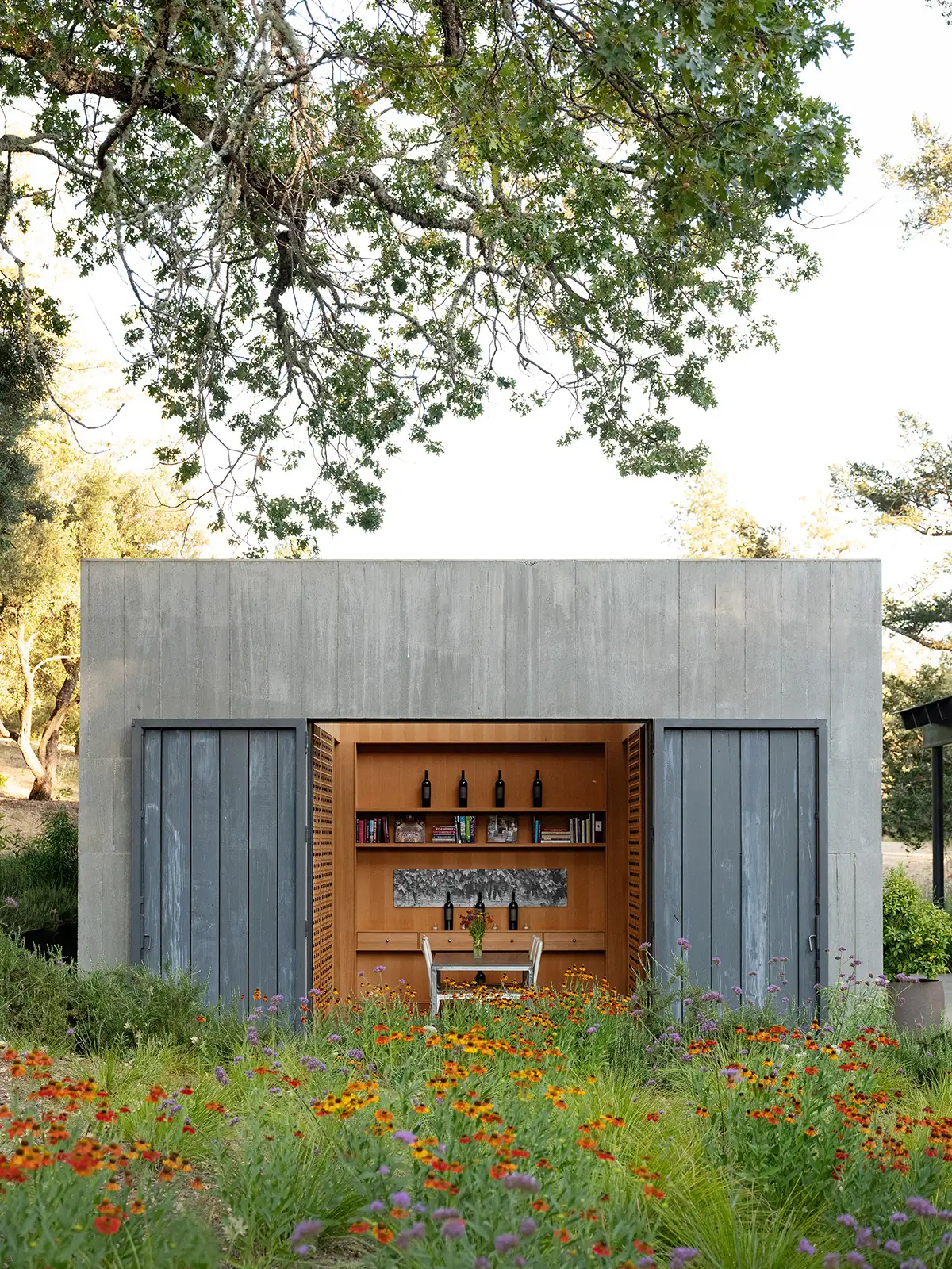 A small concrete structure with open sliding doors revealing a wooden interior shelf with wine bottles and books, positioned in a meadow of wildflowers.