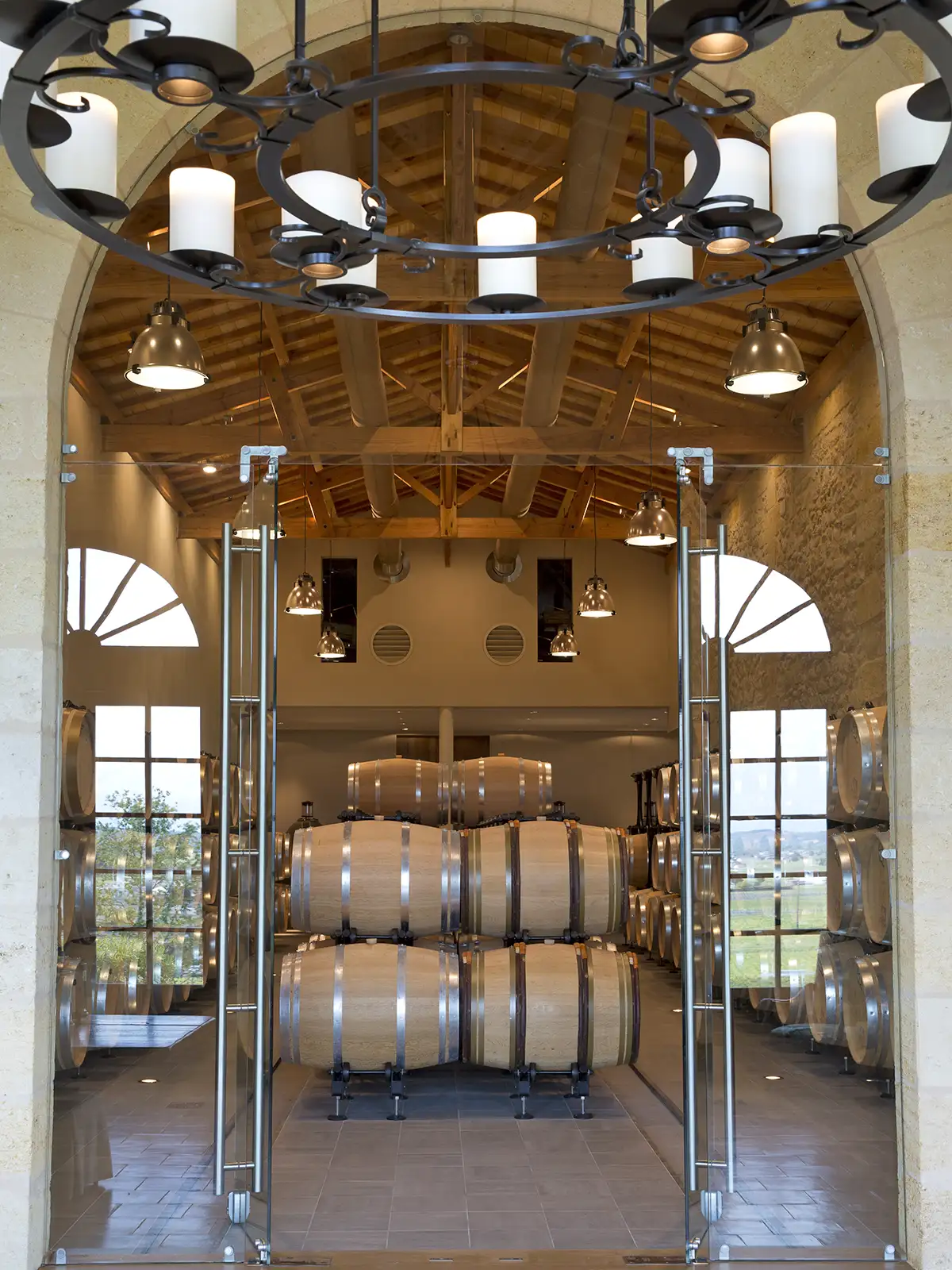 A view through glass double doors into a wine cellar lined with stacked oak barrels, featuring a large circular chandelier in the foreground.