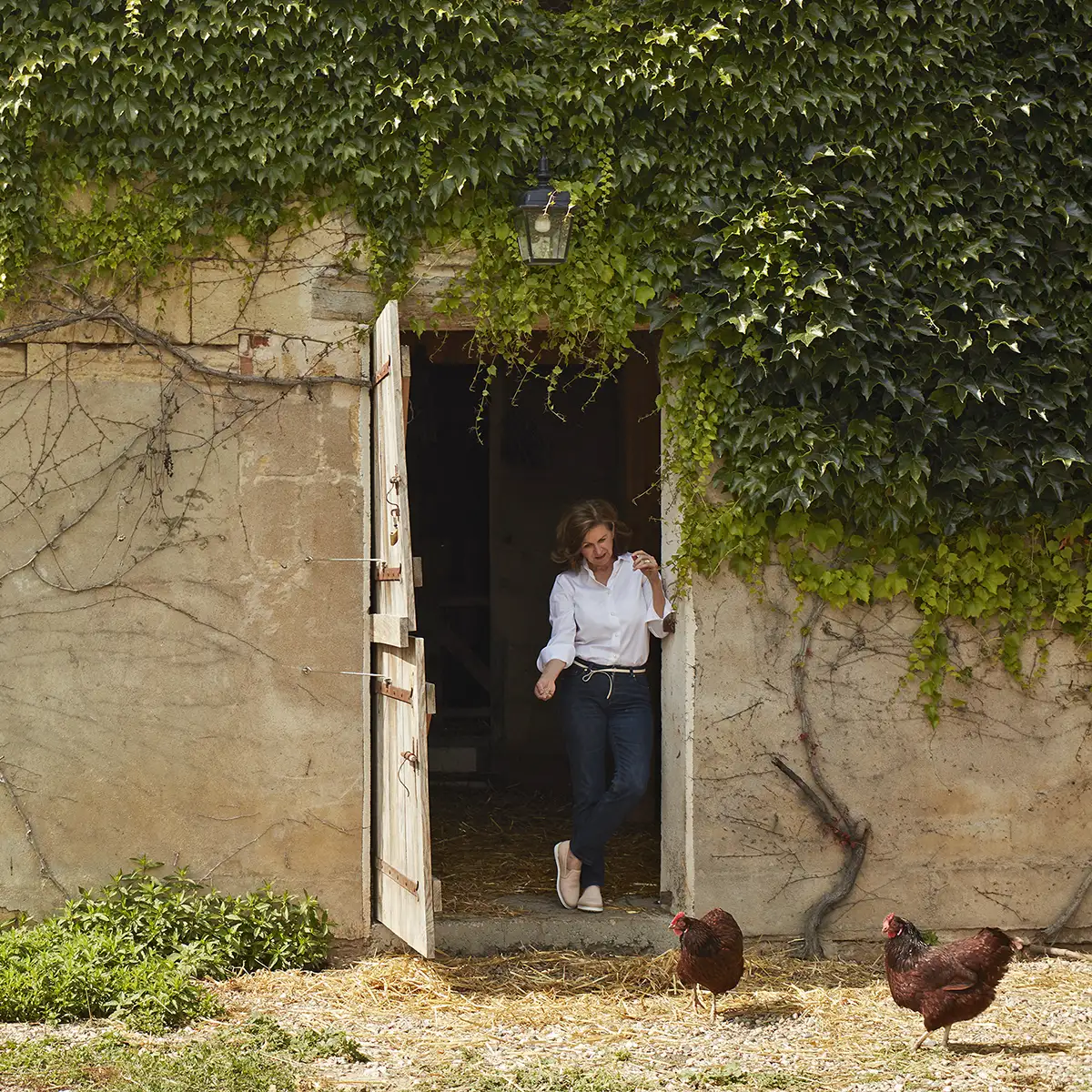 A woman in a white shirt and jeans stands in the doorway of an ivy-covered stone building with two chickens nearby.