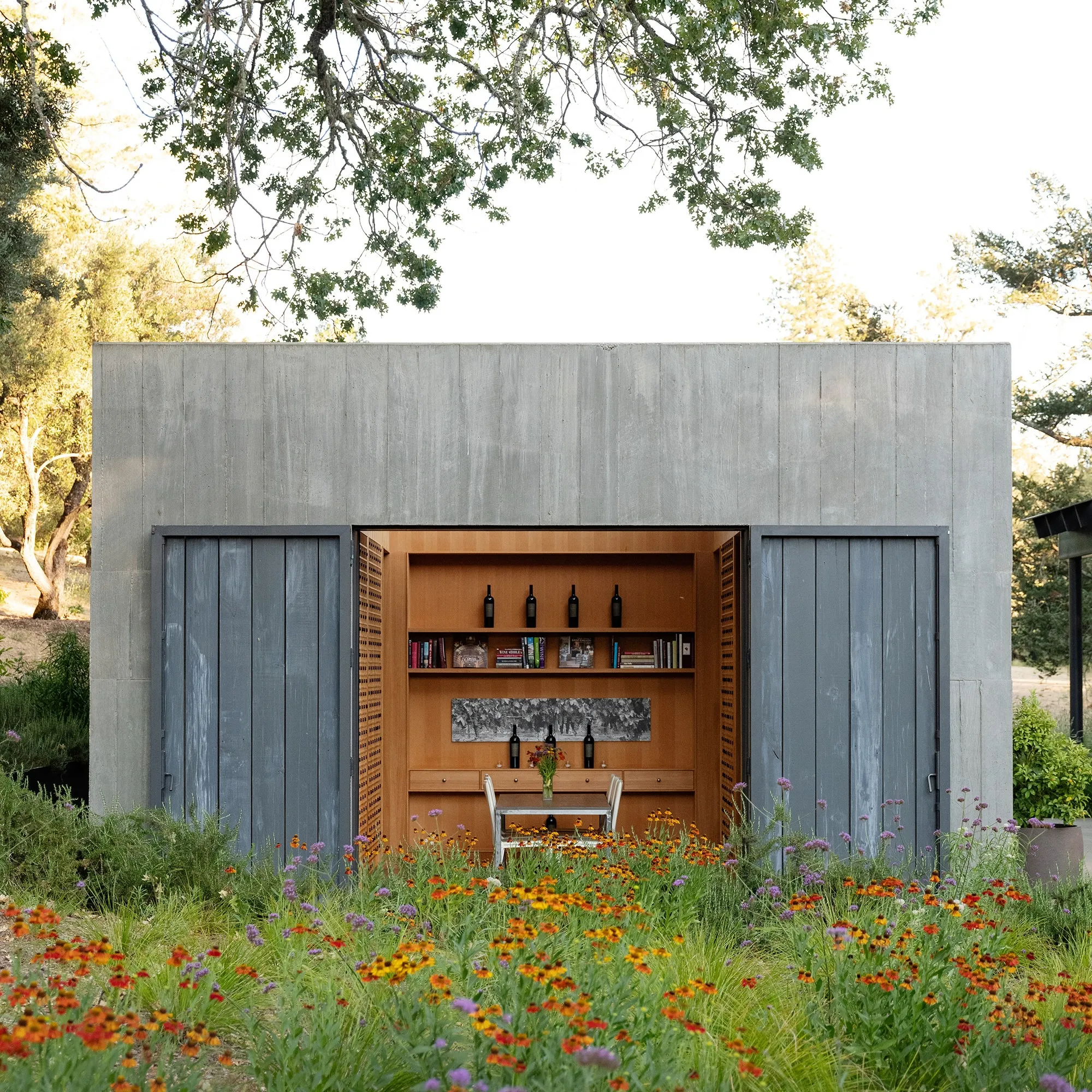 A concrete building with sliding doors open to reveal an interior wall of wooden shelves holding wine bottles and books, positioned behind a small table and chairs in a field of wildflowers.